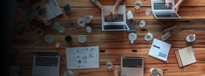 Top-down view of a team collaborating at a wooden table using laptops and tablets, with coffee cups and devices arranged neatly in a modern, digital-first workspace.