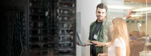 Two IT professionals stand near a server room, discussing technical details while reviewing information on a laptop in a modern office environment.