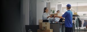 Retail store staff handing packaged orders to a delivery worker at a checkout counter, illustrating order fulfillment and last-mile delivery in a retail environment.