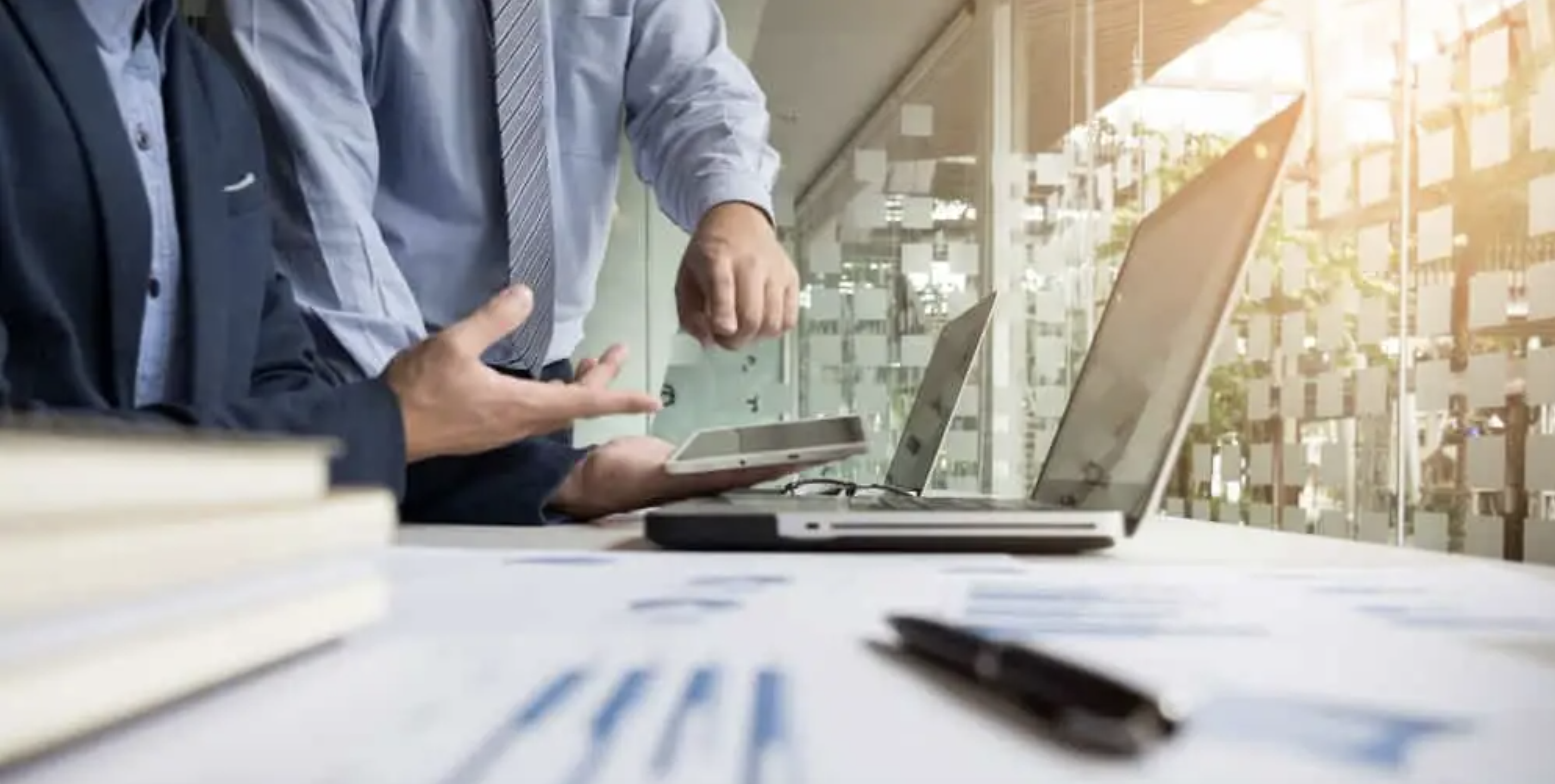Two business professionals collaborate at a desk with laptops and a tablet, reviewing data and discussing cloud-based workflows in a modern office setting.