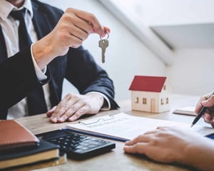 A real estate agent hands over a house key across a desk during a property transaction, with a small model home, contract paperwork, and a pen visible, symbolizing home buying, leasing, or real estate services.