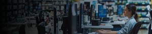 A retail employee sits at a workstation with multiple computer monitors, focused on system screens in a store environment, representing back-office retail operations, inventory management, and point-of-sale support.